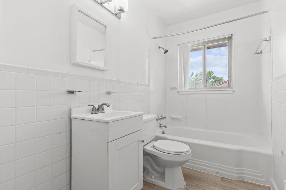 Clean white bathroom with tile walls, vanity, toilet, and bathtub next to a bright window.