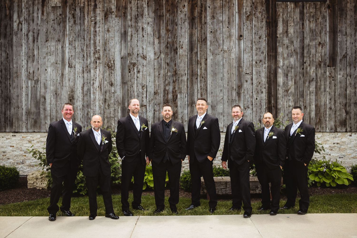 Groom and groomsmen in black tuxedos posing in front of a rustic wooden barn wall at a wedding.