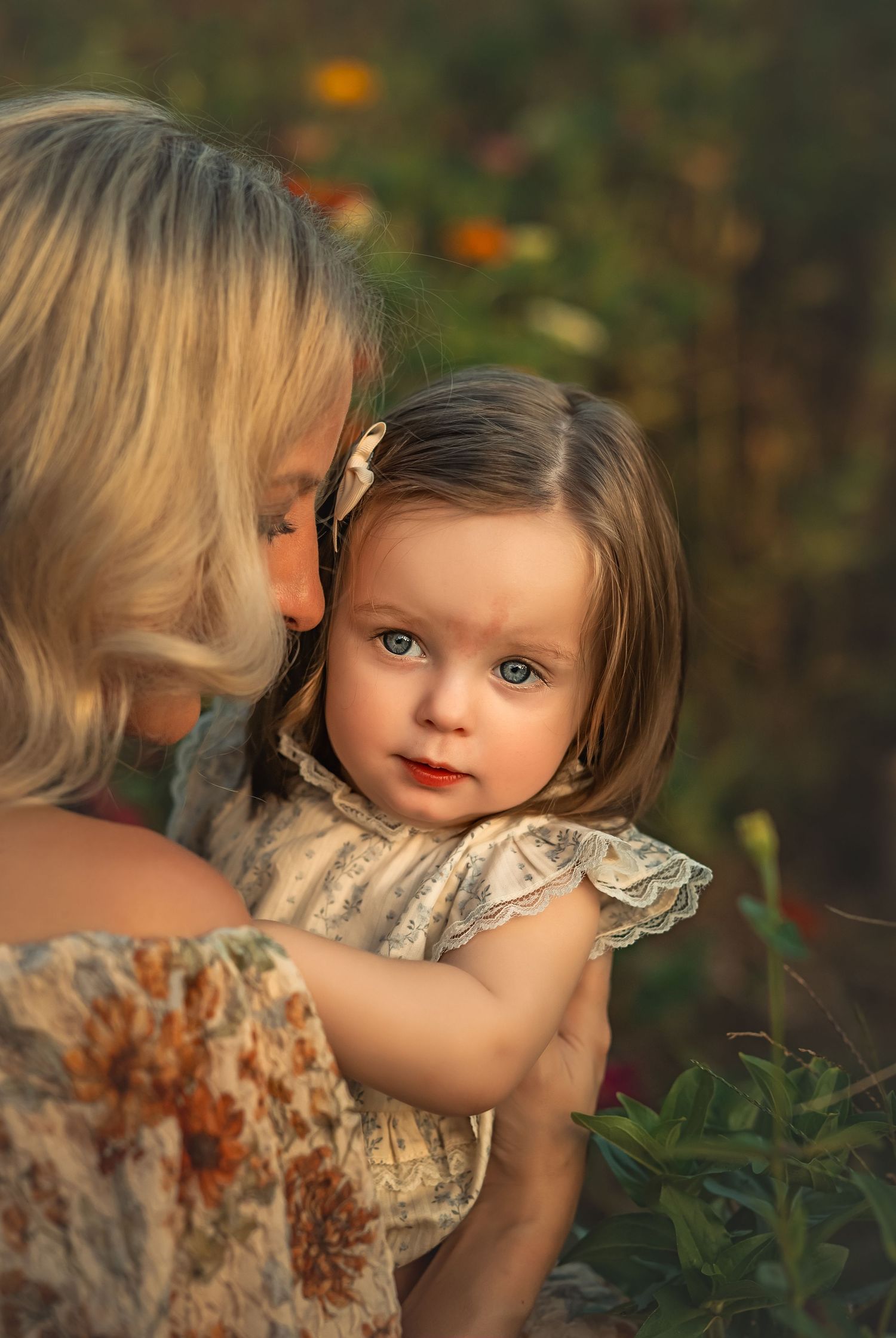 A blonde woman tenderly kisses a toddler girl with blue eyes in a sunlit garden setting.