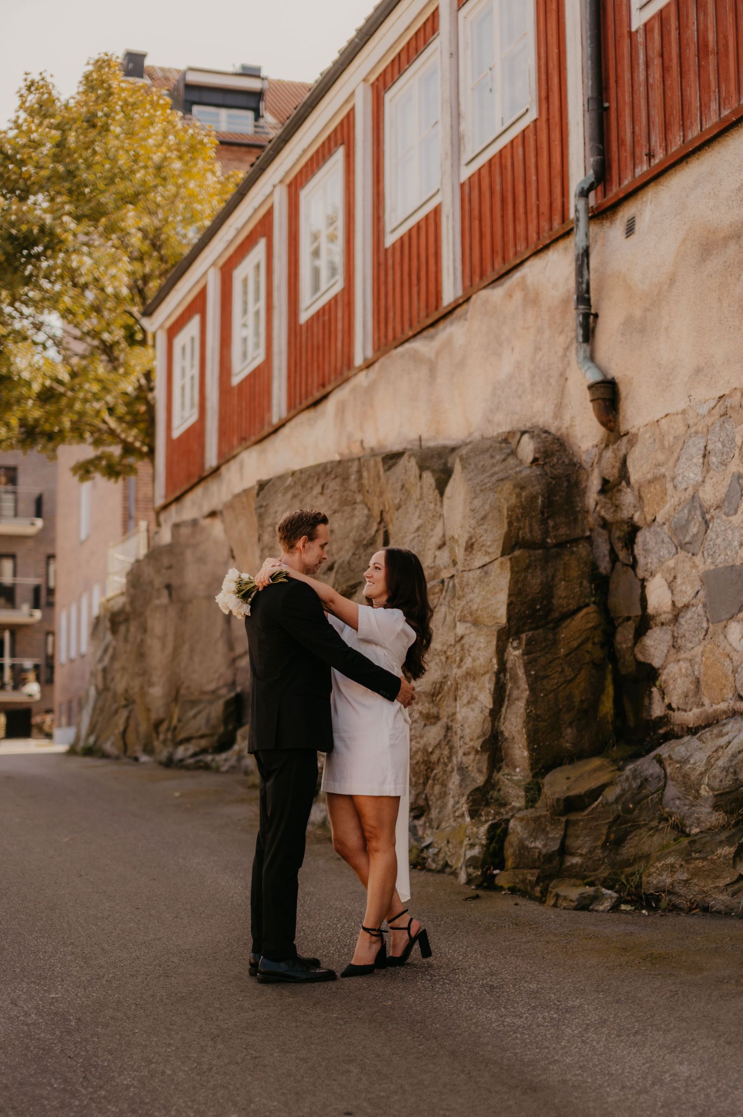 Couple in wedding attire holding hands on a cobblestone street beside a red building and stone wall.