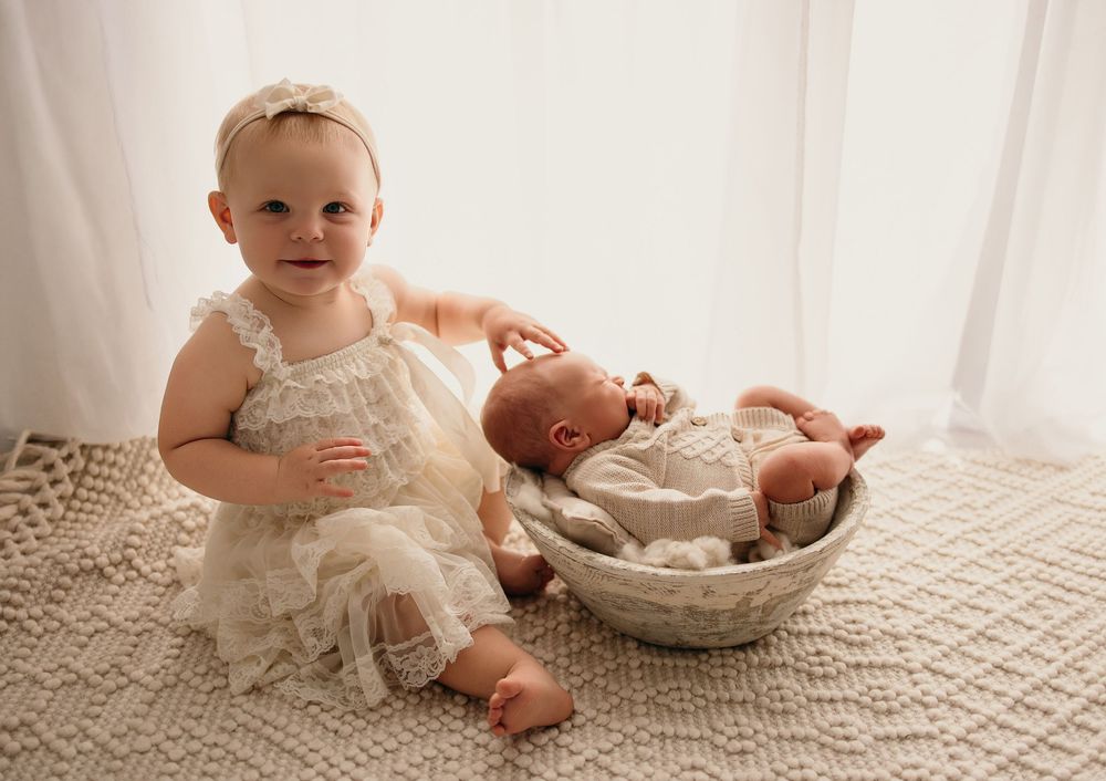 Toddler girl in lace dress sits beside newborn baby in woven bowl on cream knit blanket in a Bettendorf natural light studio.