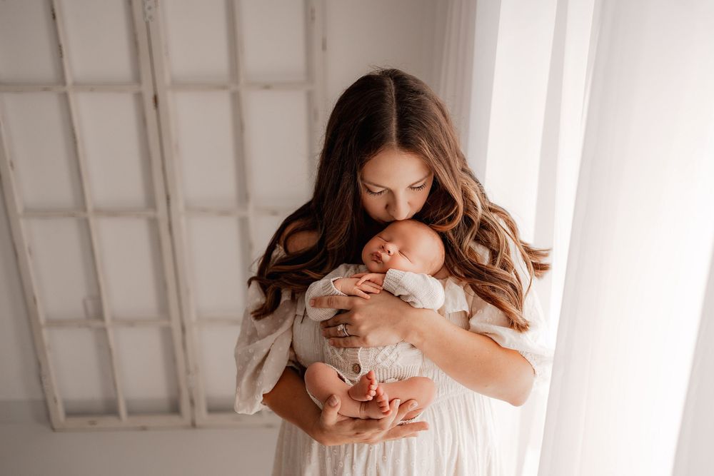 A mother in a white dress tenderly kisses her sleeping newborn baby during her newborn session in a Bettendorf photo studio.