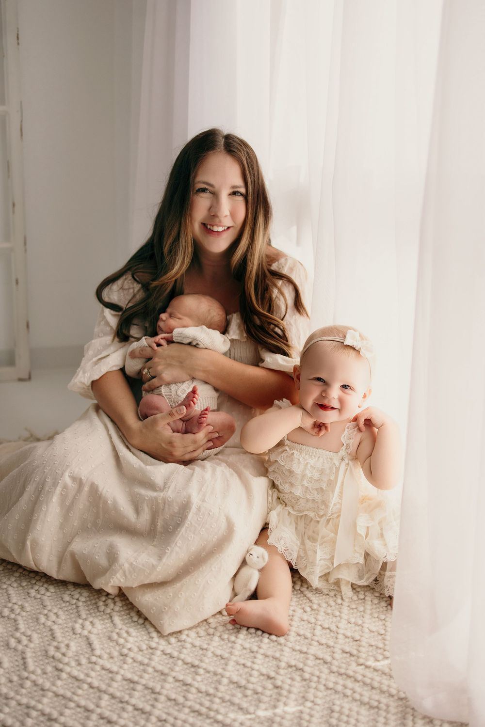 A smiling mother in a cream dress sits by a window holding a newborn - Bettendorf Newborn Photographer