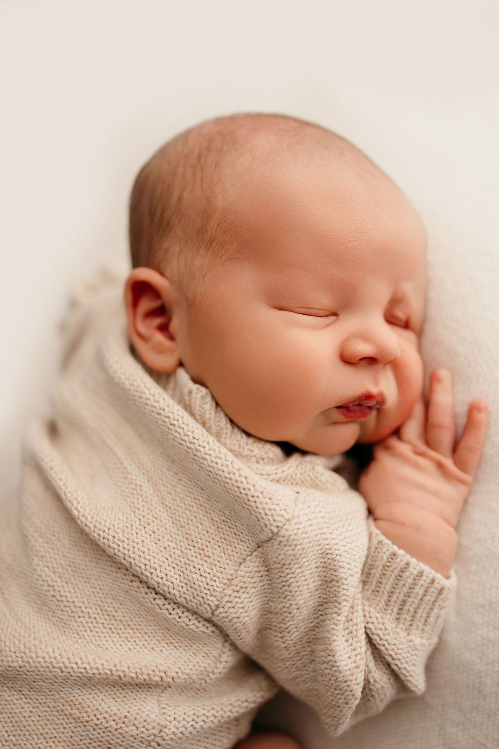 Peaceful newborn baby sleeping in a cozy beige knit sweater on a soft white background for a newborn session in Bettendorf Iowa.