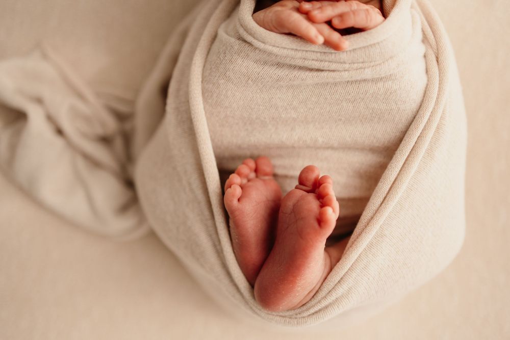 Close-up of newborn baby feet peeking out of a soft beige swaddle wrap on a neutral background in Bettendorf Newborn Studio.