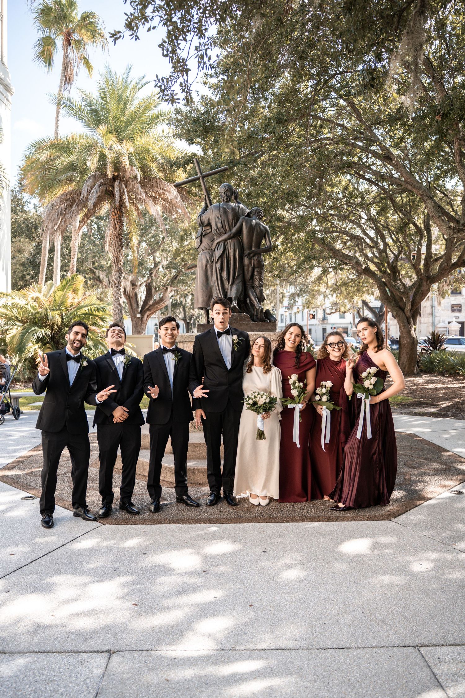 Wedding party poses before a statue outdoors, groomsmen in tuxedos and bridesmaids in burgundy gowns with bride in white in St Augustine FL