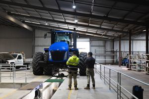 Two workers inspecting a large blue New Holland tractor inside a modern farm machinery shed.