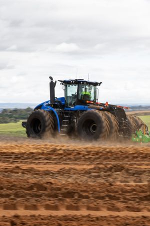 Large blue New Holland tractor pulling cultivation equipment across a dusty plowed farm field.
