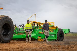 Two workers in hi-vis gear standing beside green agricultural spray equipment on a dirt field.