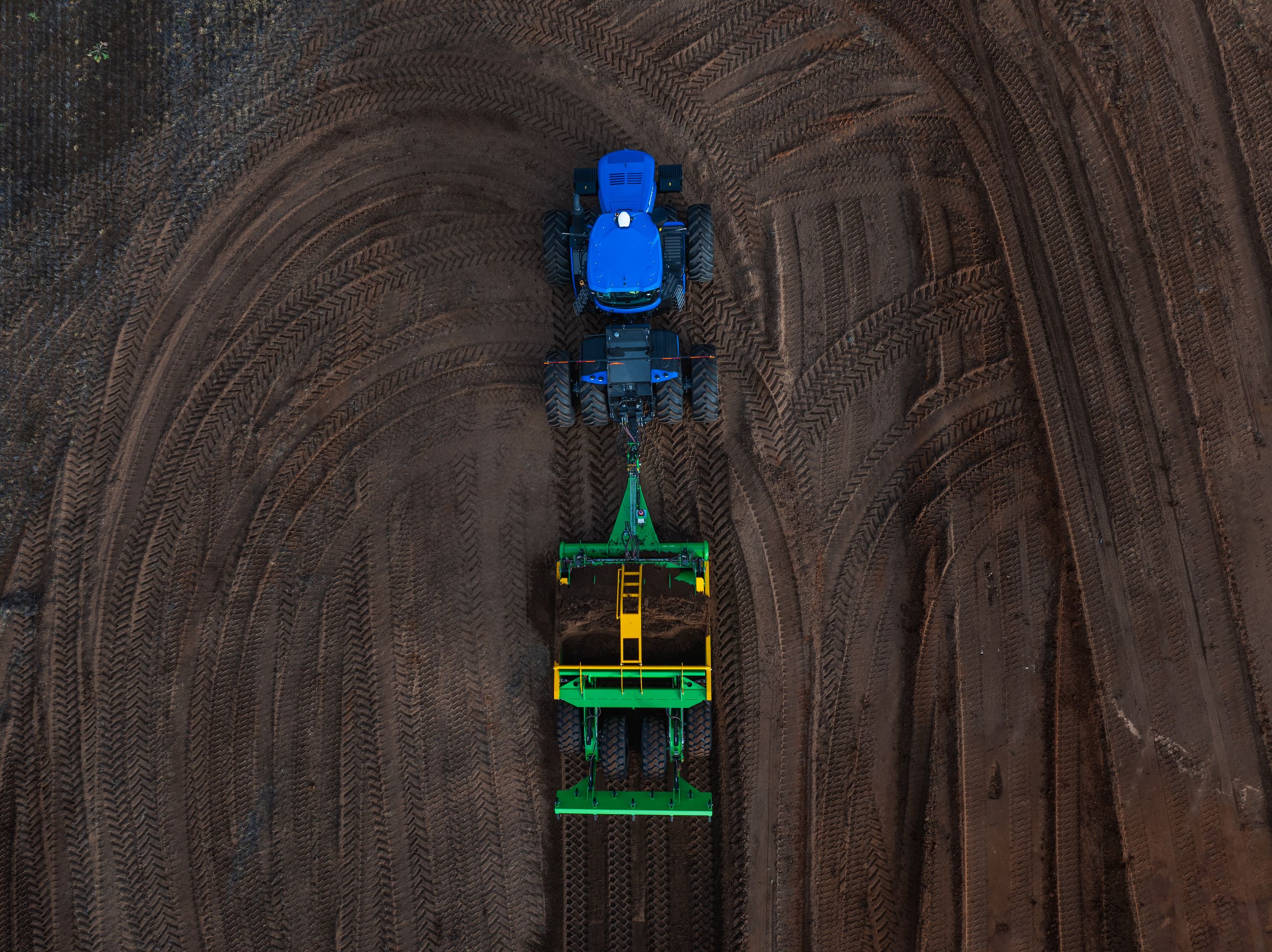 Aerial view of a blue tractor with green farming equipment cultivating a large agricultural field.