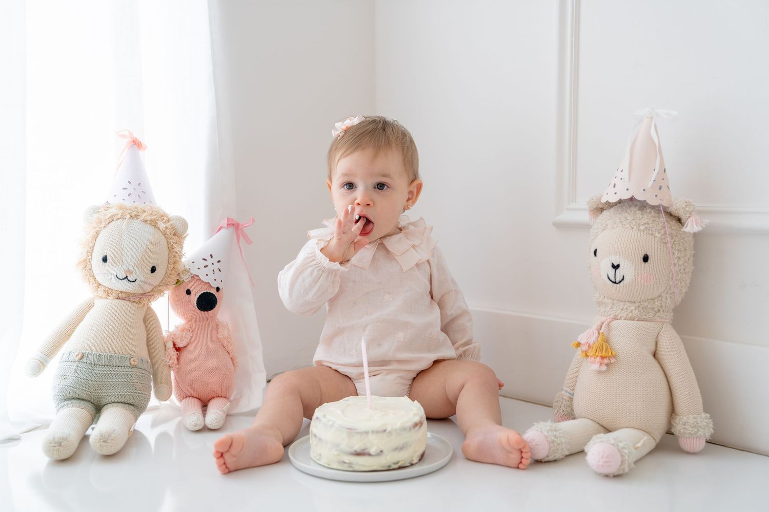 Cute baby celebrating first birthday sitting with stuffed animal toys beside a small white smash cake.