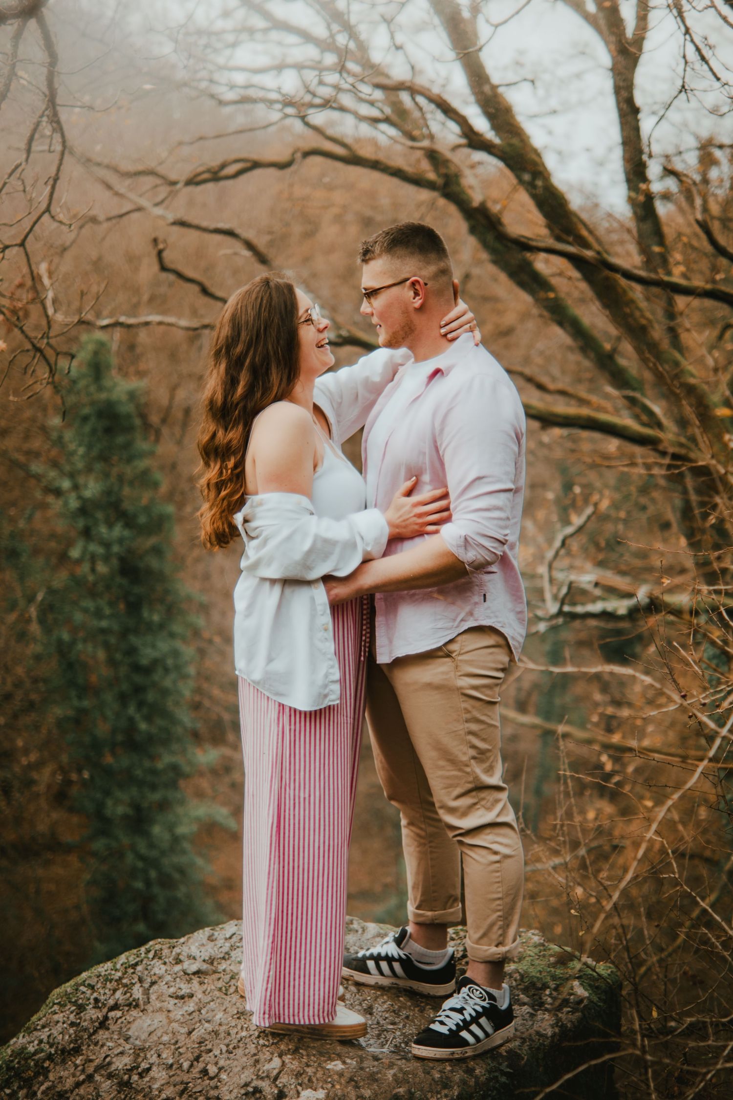 Couple embracing on a rock in an autumn forest, wearing casual outfits, sharing a romantic moment outdoors.