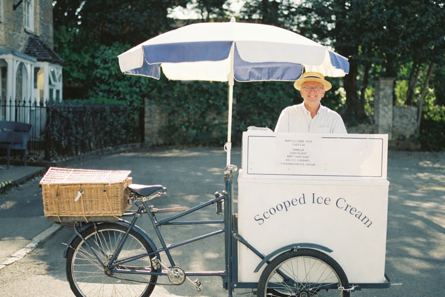 Man in straw hat selling scooped ice cream from a vintage bicycle cart with blue and white umbrella outdoors.