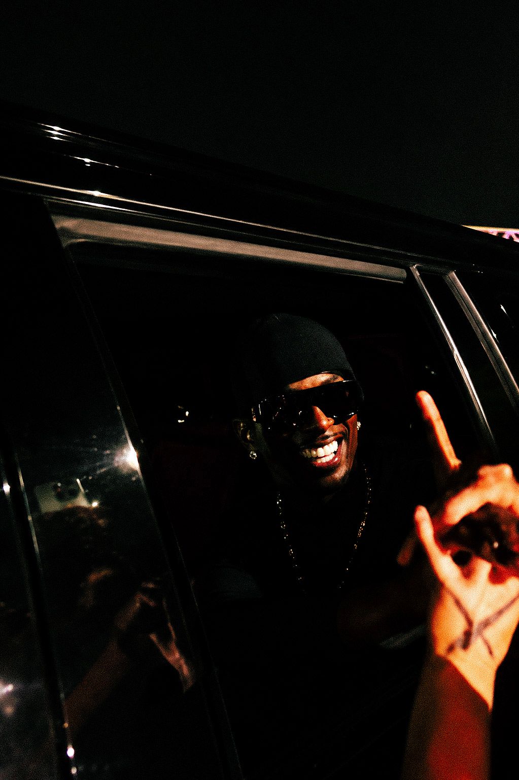 Smiling man wearing sunglasses and black cap leaning out of a car window at night making hand gestures.