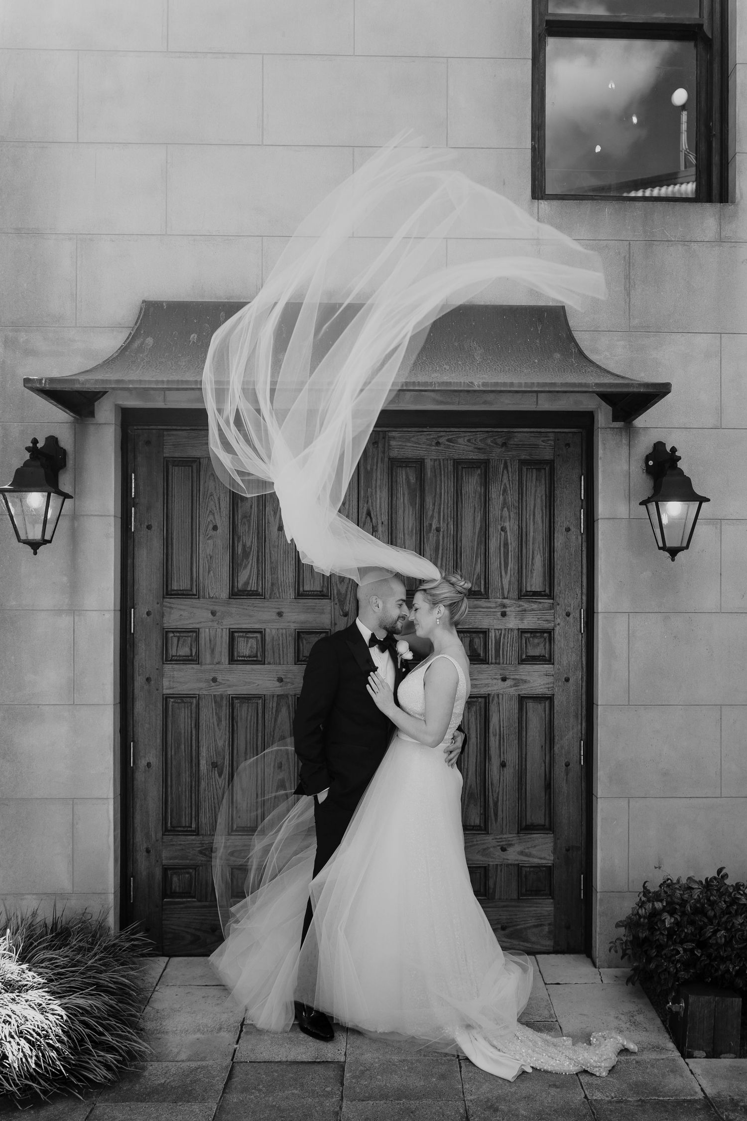 Black and white photo of bride and groom kissing as veil flows dramatically in wind before elegant wooden doors.