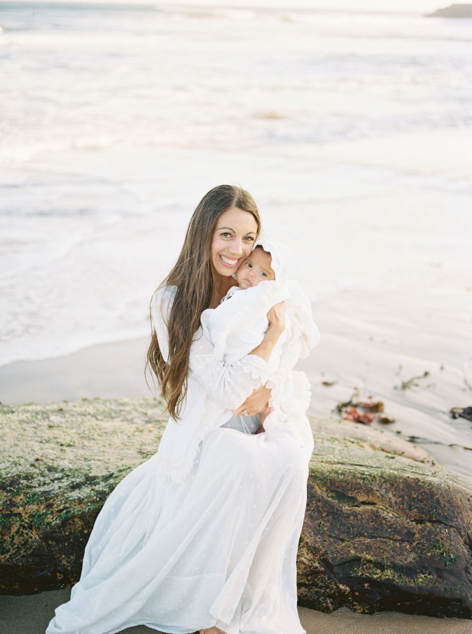 Woman in white dress holding baby on rocky beach with ocean waves in background.