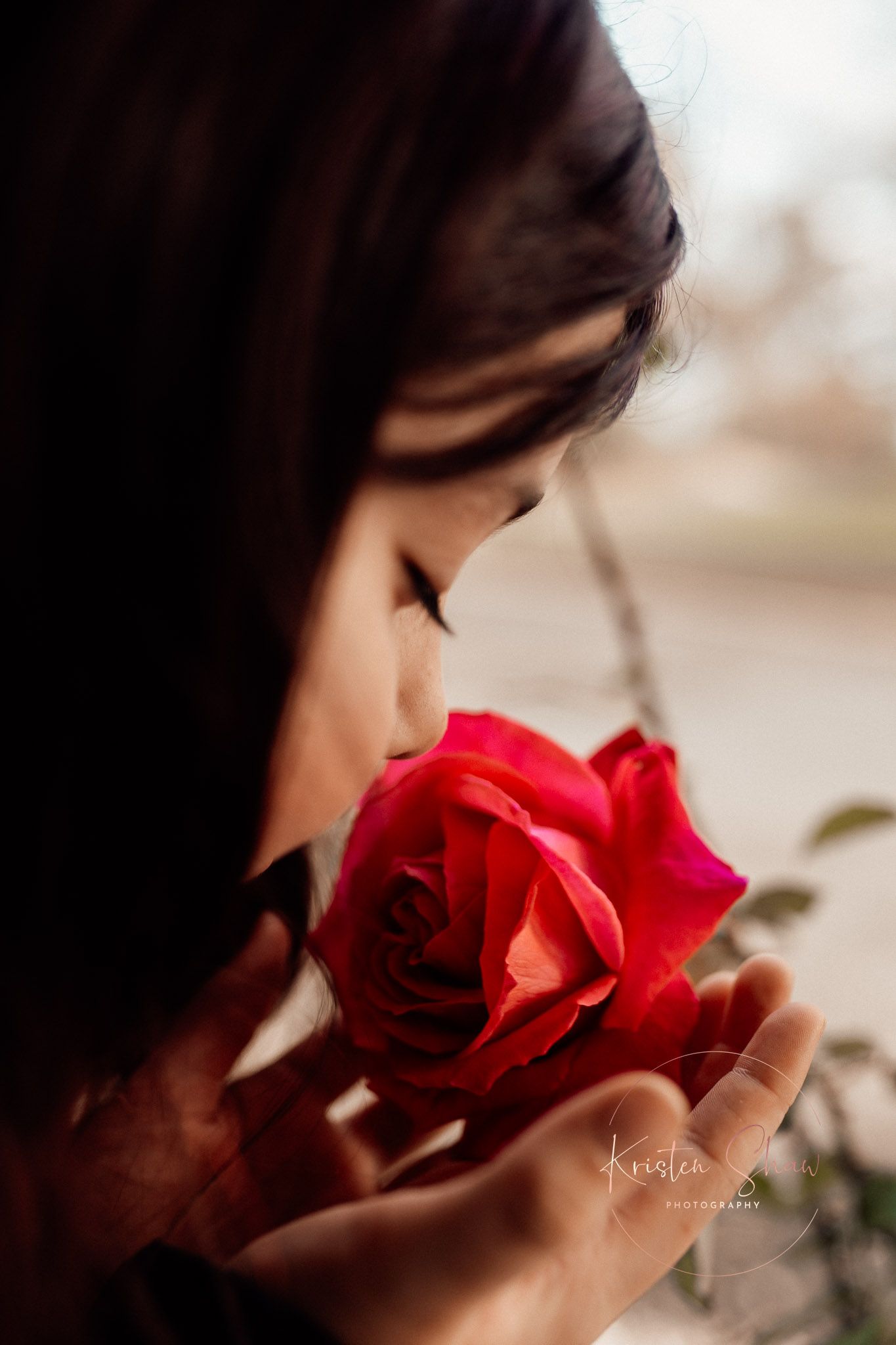 A young woman with dark hair smells a vibrant red rose held in her hands in a moody, softly lit portrait.