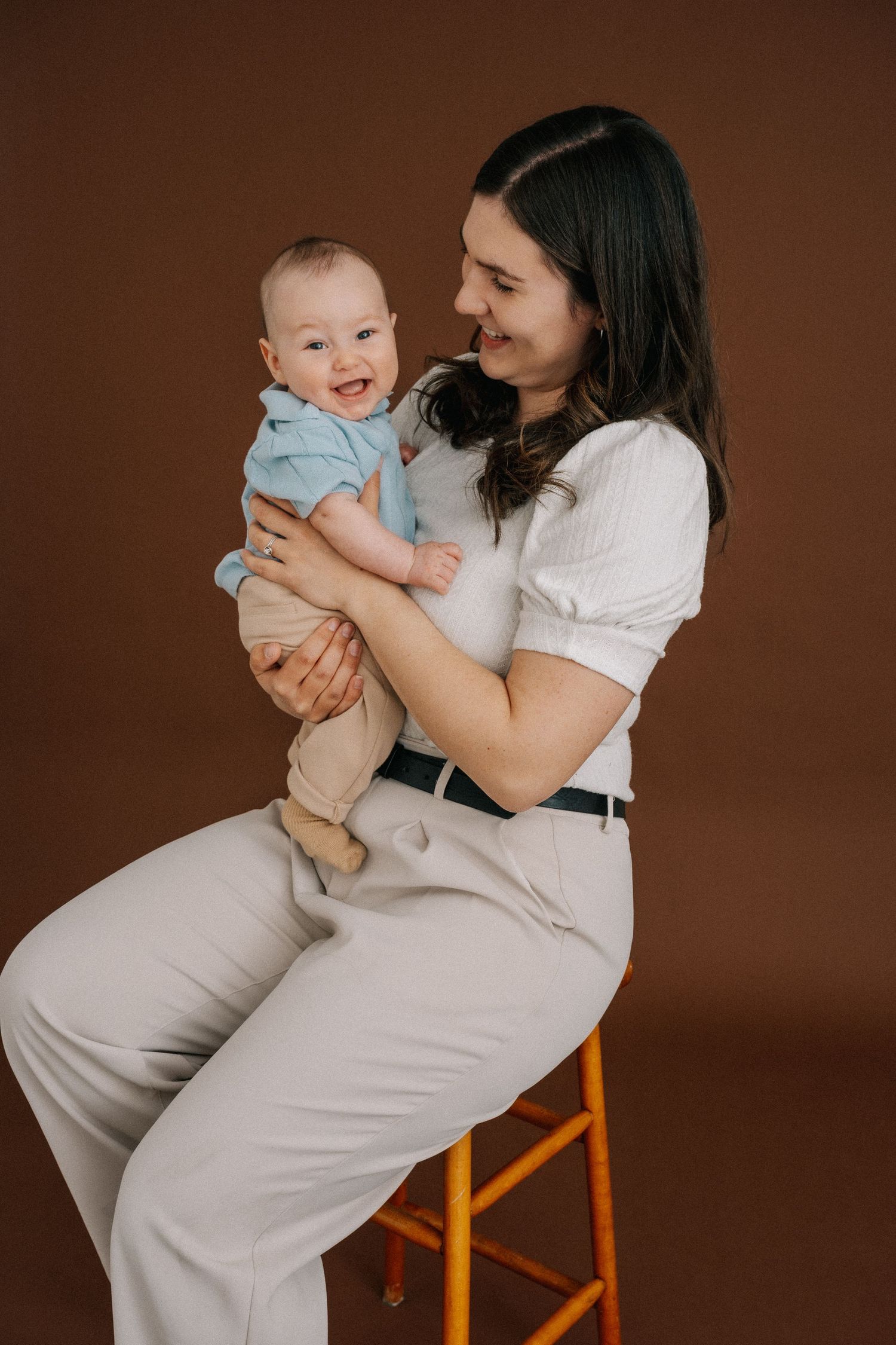 A smiling mother sits on a stool holding her happy baby dressed in blue against a brown background.