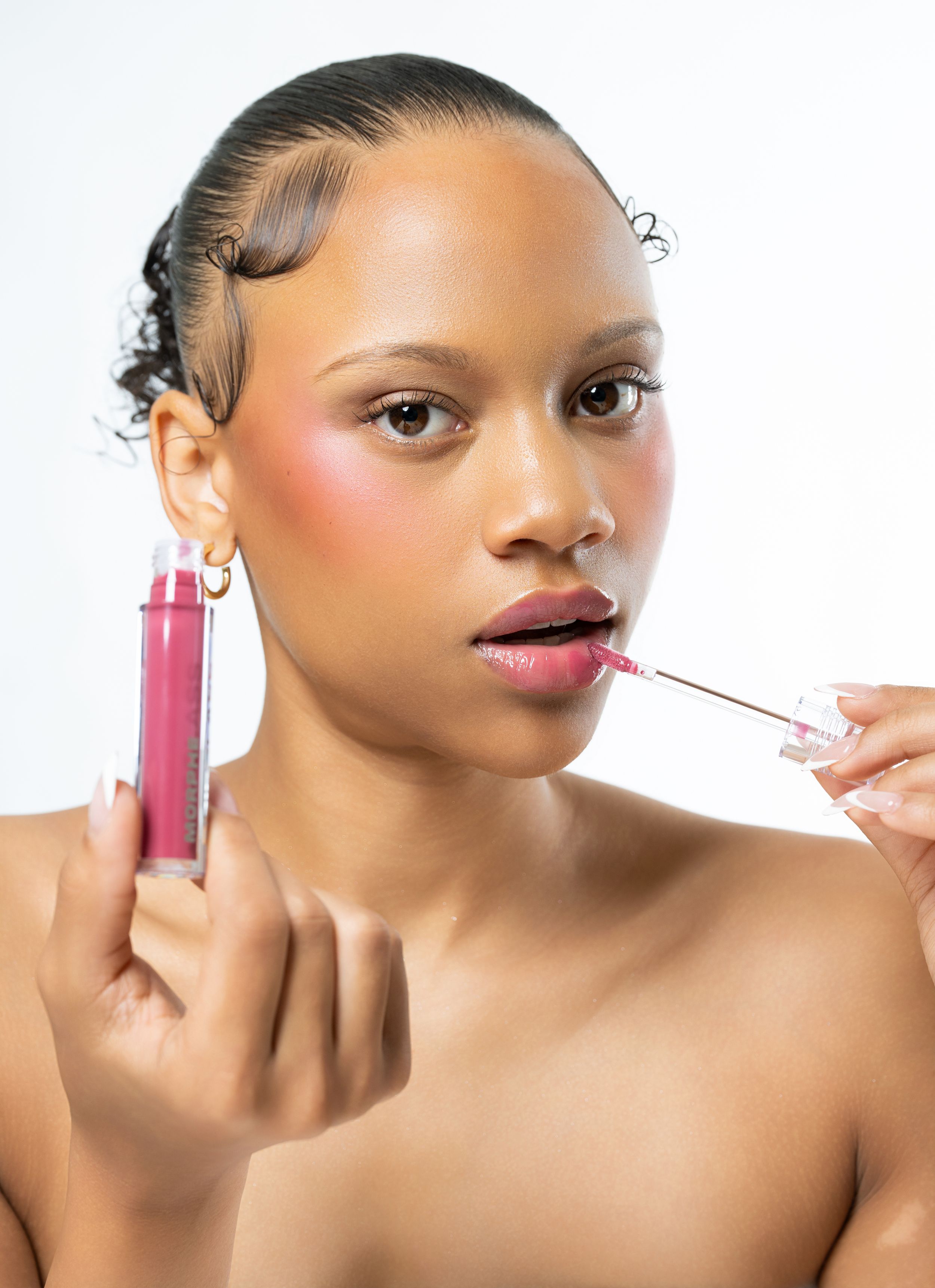 Woman applying pink lip gloss with wand applicator, holding bottle, wearing gold hoop earrings, beauty product photo.