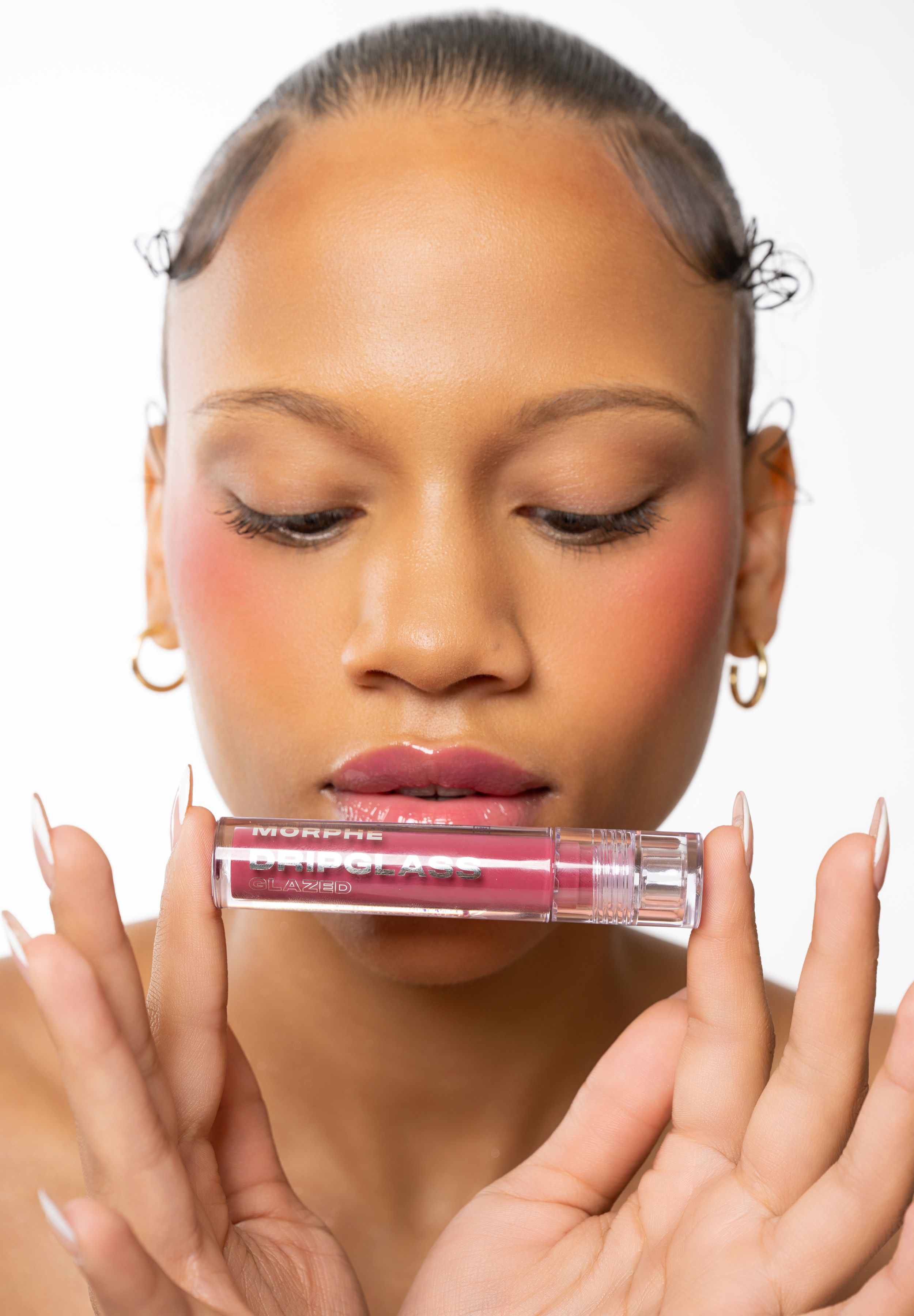 Woman holding a Morphe Drip Gloss lip gloss to her lips, wearing gold hoop earrings and a rosy blush makeup look.