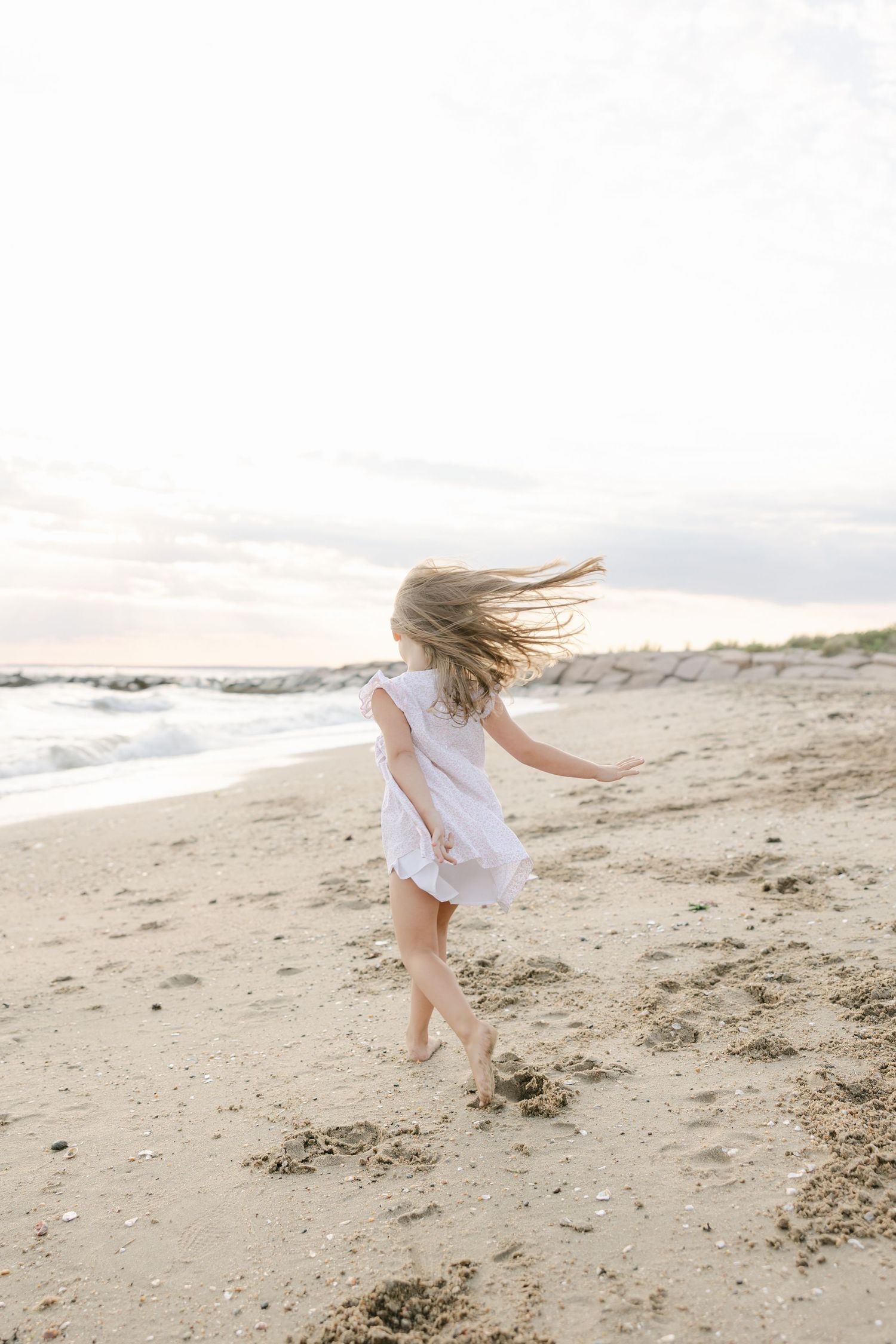 A young girl in a white dress runs barefoot along a sandy beach, her hair blowing in the wind.