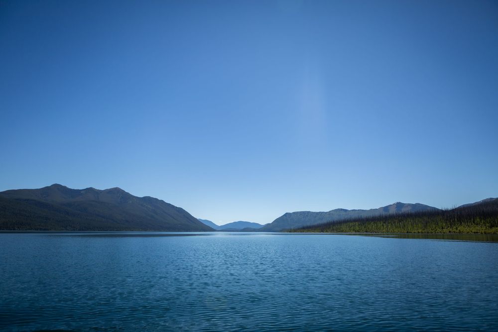 Serene blue mountain lake with calm waters, forested shores, and distant peaks under a clear sky.