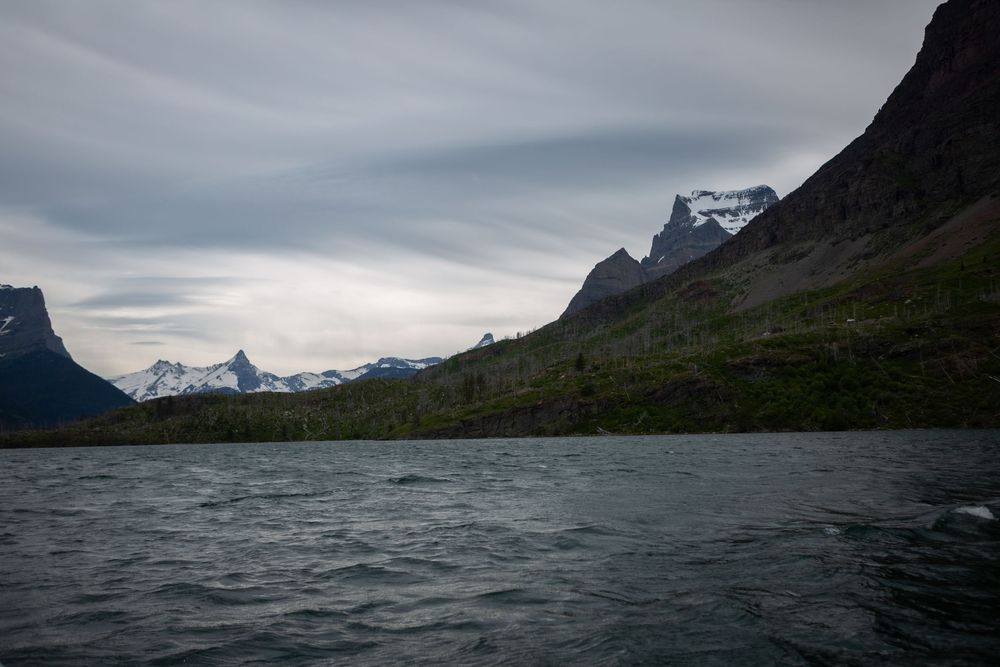 Dramatic mountain lake landscape with snow-capped peaks, rocky cliffs, and evergreen forests under cloudy skies.