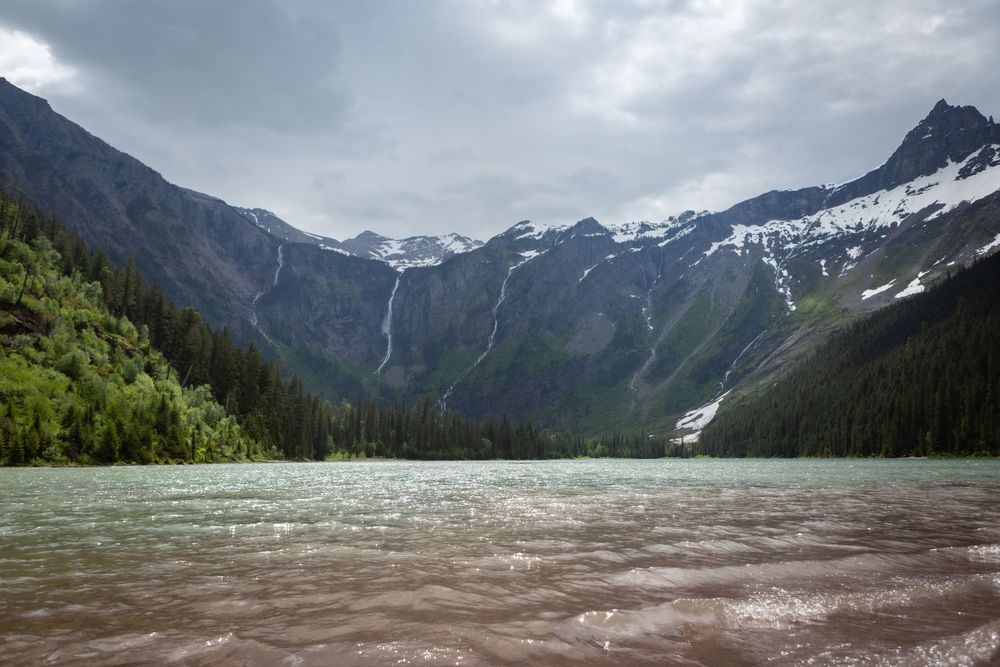 Turquoise glacial lake with choppy water, surrounded by snow-capped mountains and dense evergreen forests under cloudy skies.