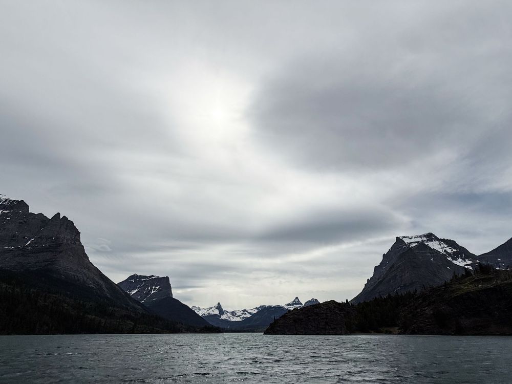 Dramatic mountain landscape with snow-capped peaks reflecting over a calm lake under moody overcast skies.