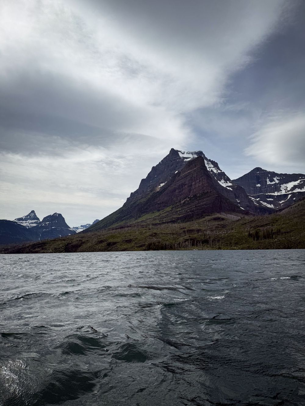 Choppy glacial lake waters with dramatic snow-capped mountain peaks rising under cloudy skies in Glacier National Park.