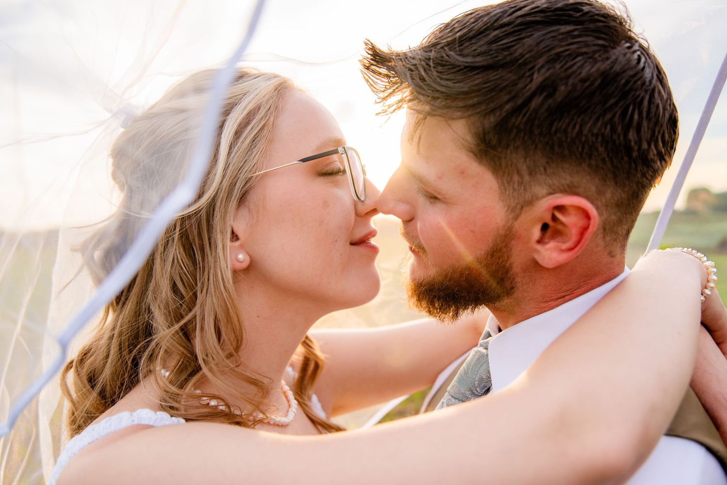 Bride and groom sharing a romantic almost-kiss under a wedding veil with golden sunset light behind them.