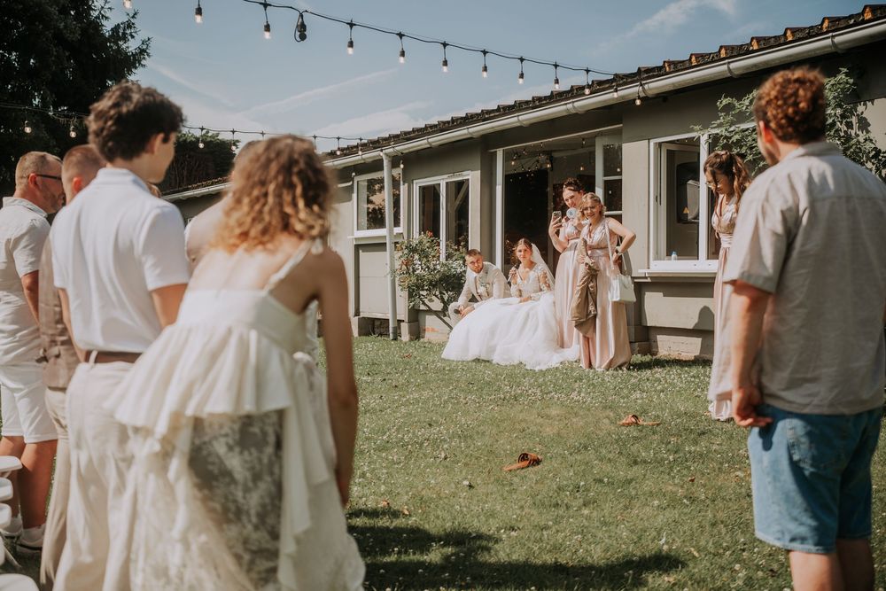 Outdoor backyard wedding ceremony with guests in white attire on green lawn, string lights, and ranch-style house backdrop.