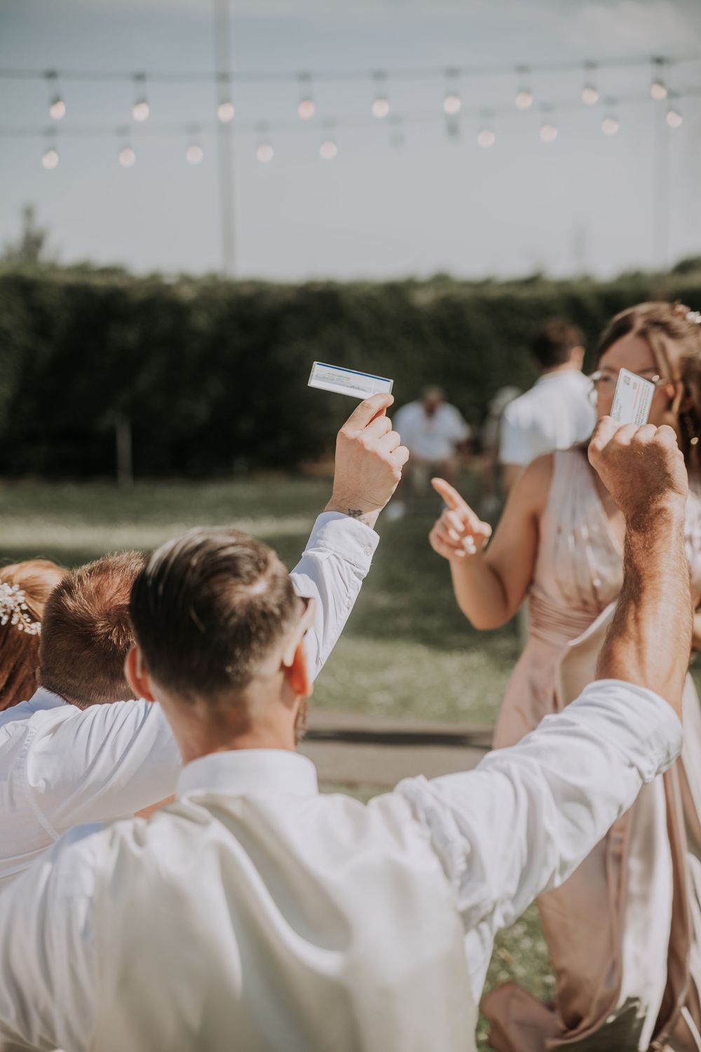 Wedding guests holding programs outdoors at a sunny garden reception with string lights and green hedges in the background.