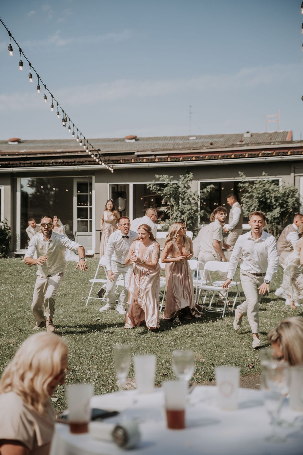 Guests in light attire joyfully run across a sunny outdoor wedding reception on a green lawn.