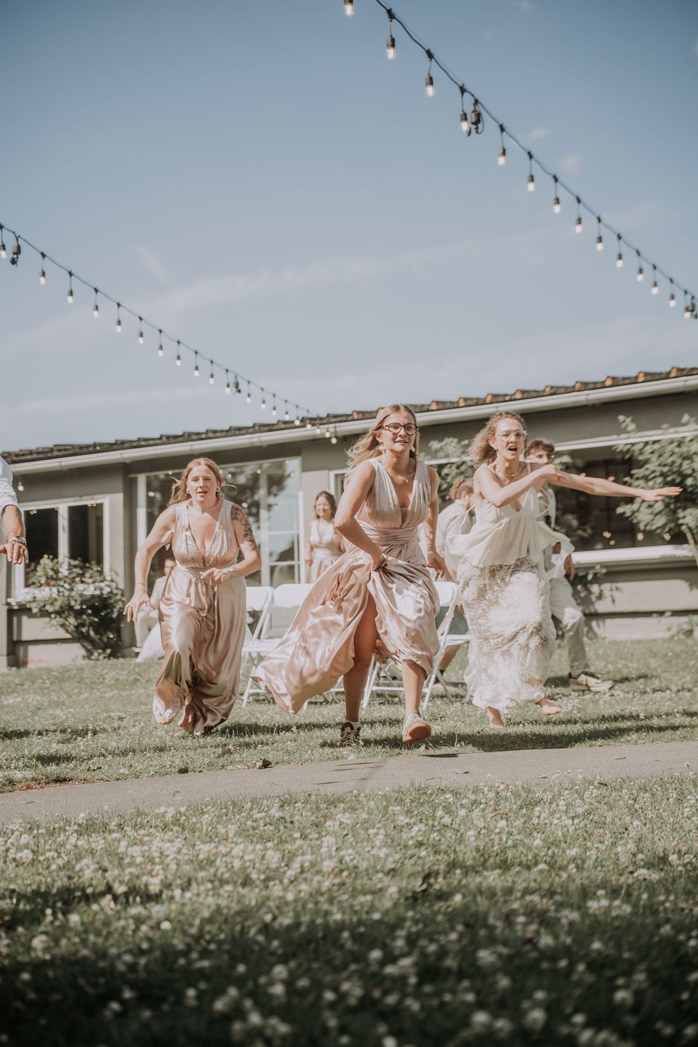 Three women in elegant dresses laughing and running outdoors at a wedding reception with string lights overhead.