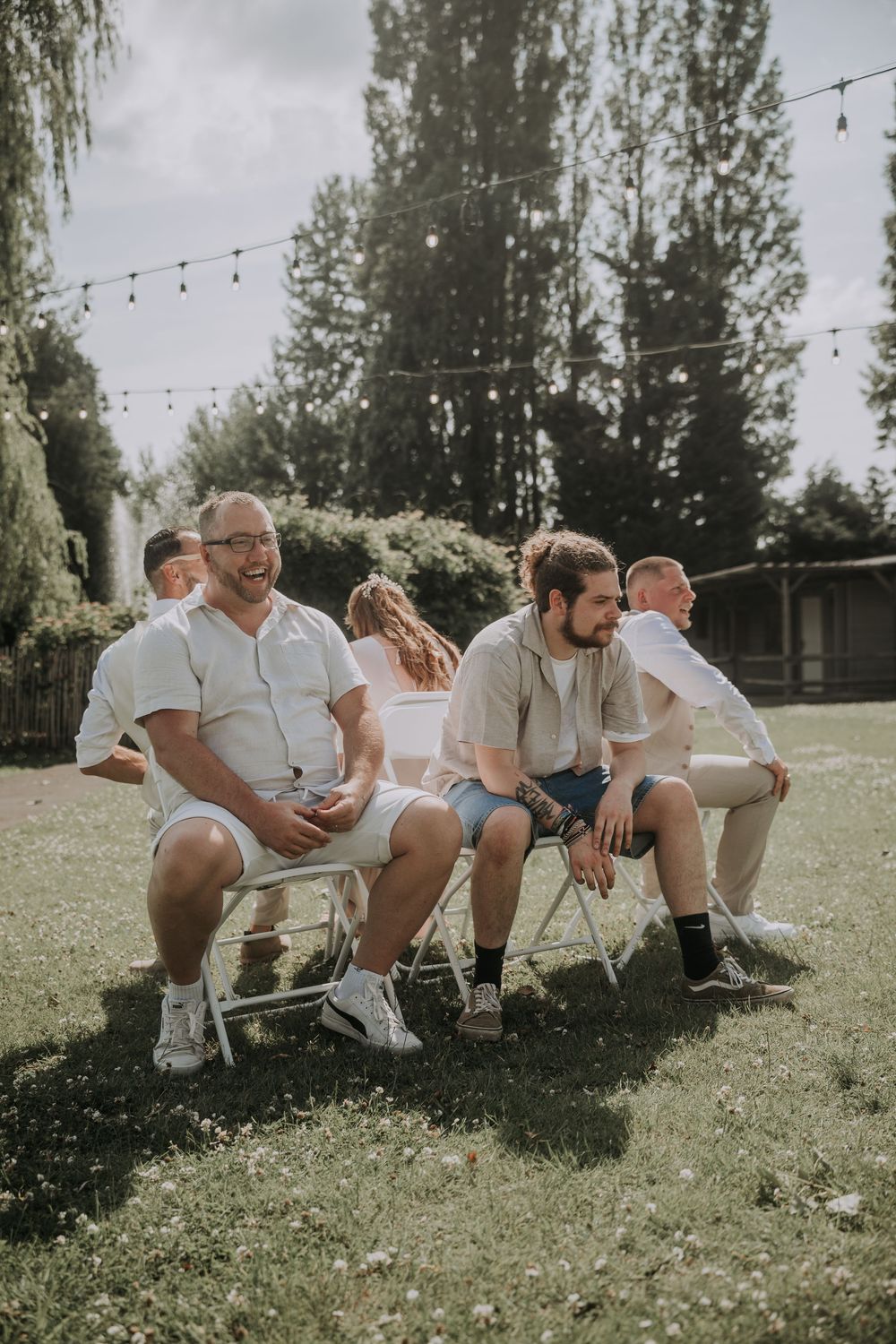 Men in casual white attire laugh and relax on folding chairs at an outdoor garden wedding celebration.