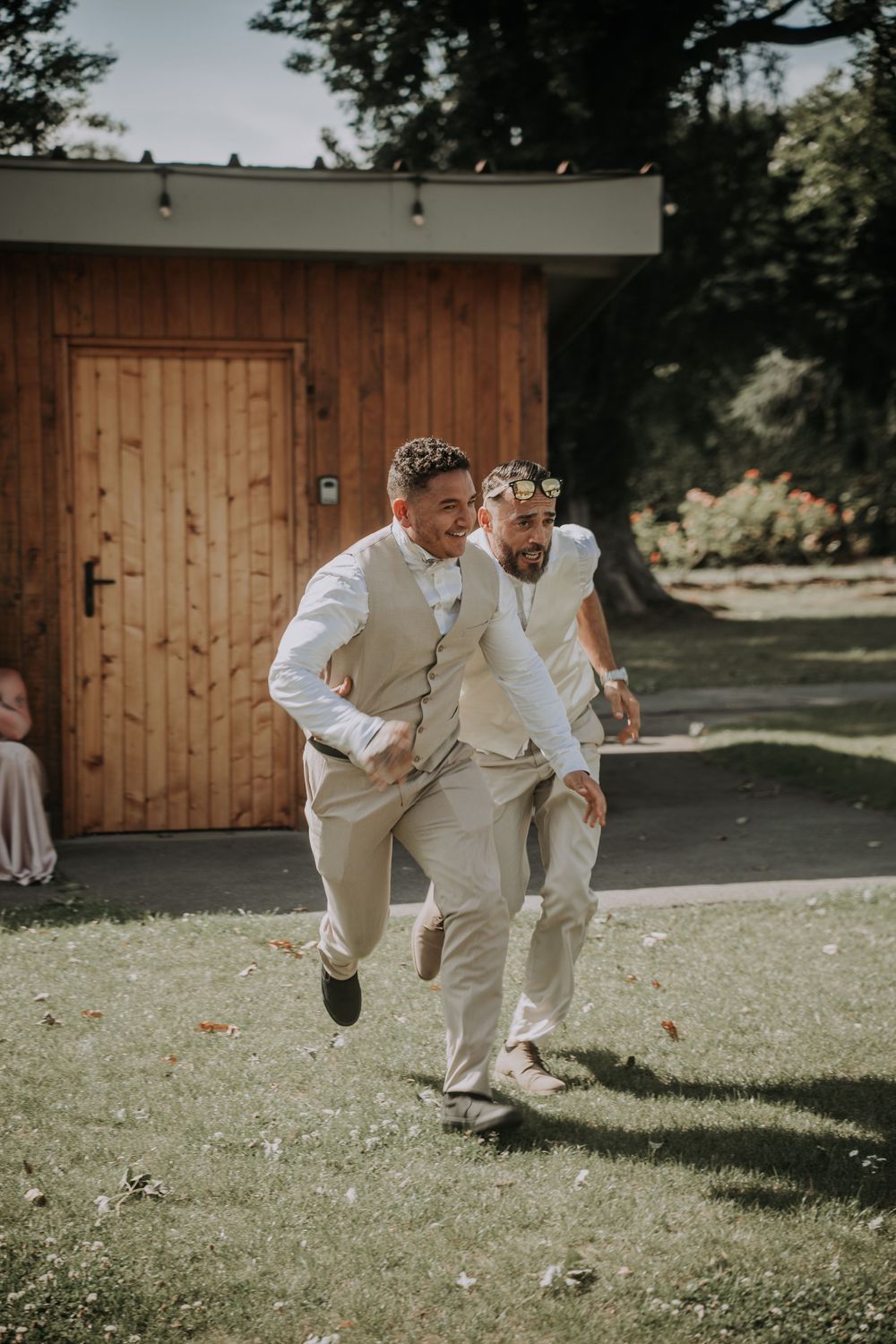 Two men in wedding attire playfully run across a sunny garden lawn during a wedding celebration.