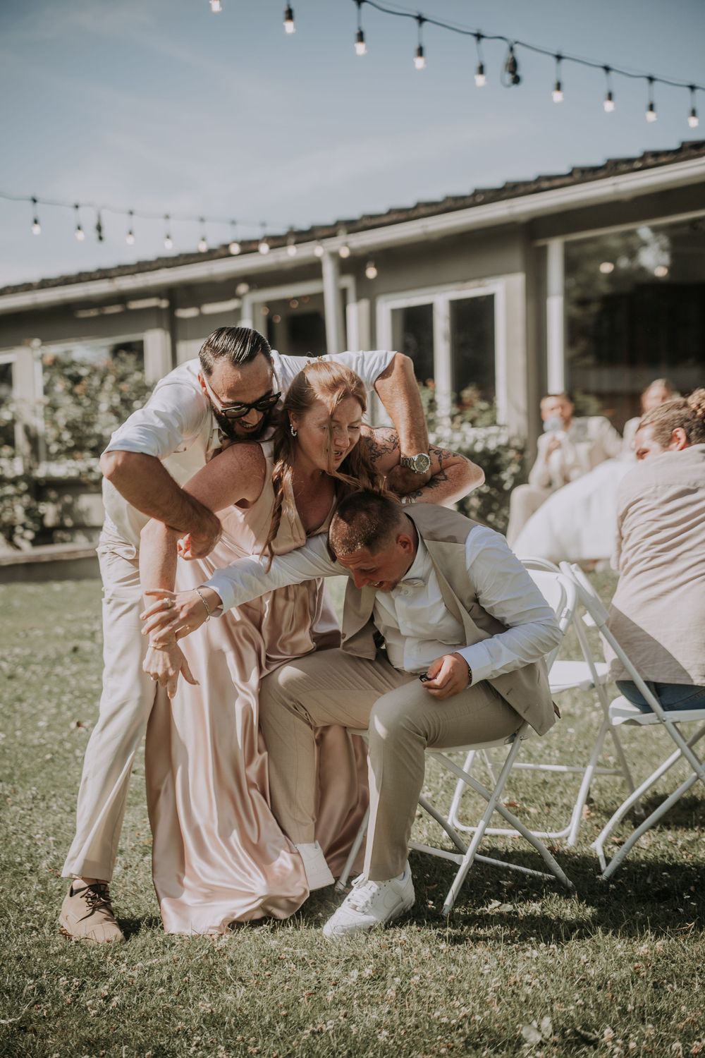Wedding guests playfully pile together on lawn during outdoor reception with string lights and white folding chairs.