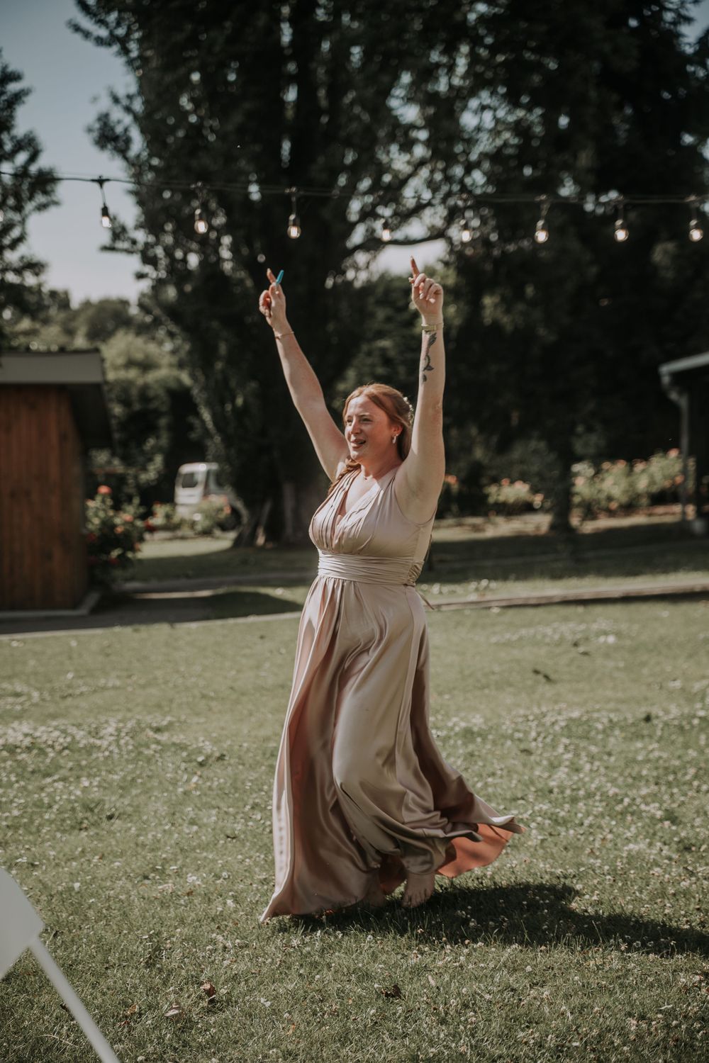 Joyful woman in flowing maxi dress raises arms triumphantly while dancing at an outdoor wedding.
