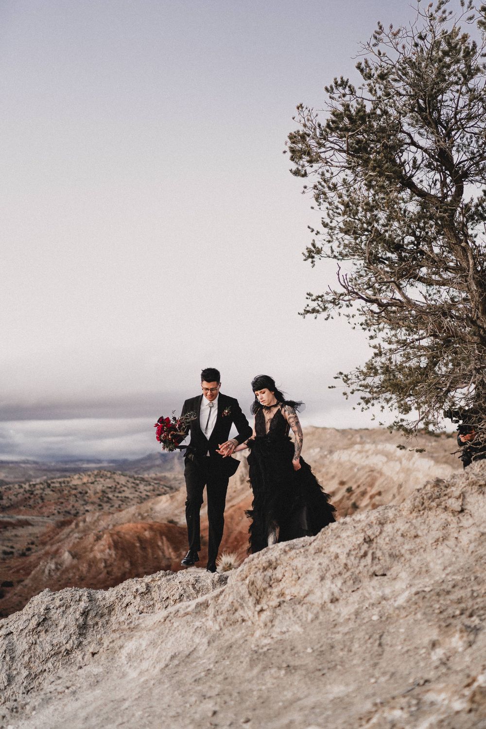 Bride in black gown and groom walk along rocky desert ridge with vast landscape and lone tree in Ojito Wilderness