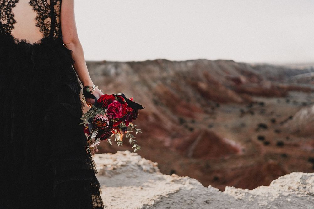 Bride in black lace gown holds deep red and burgundy bouquet against colorful desert canyon backdrop in Ojito Wilderness
