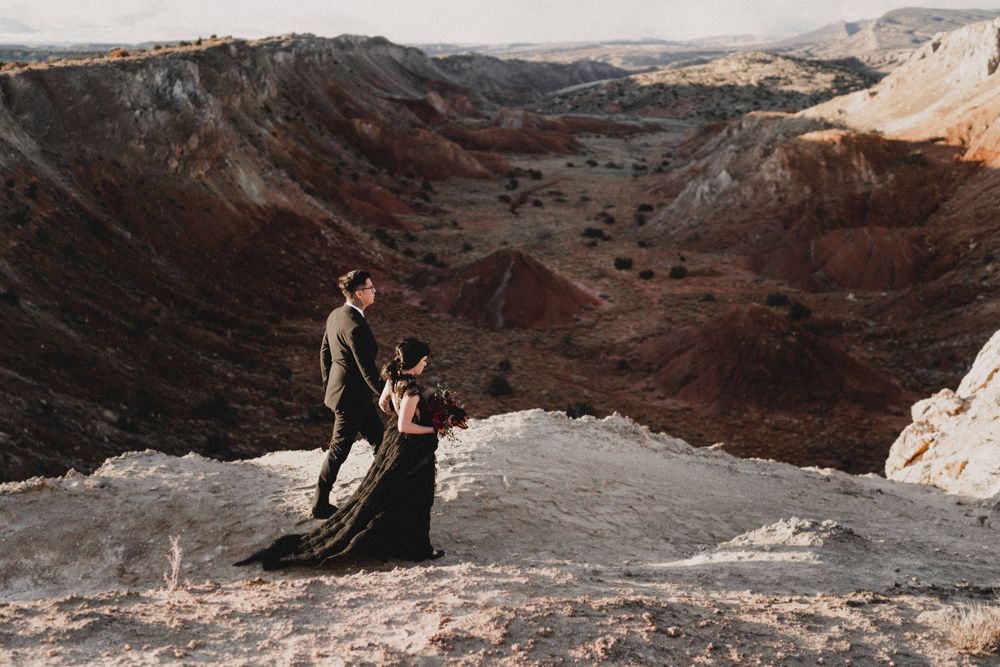 Couple in black wedding attire stands on dramatic red canyon overlook during desert elopement shoot in Ojito Wilderness