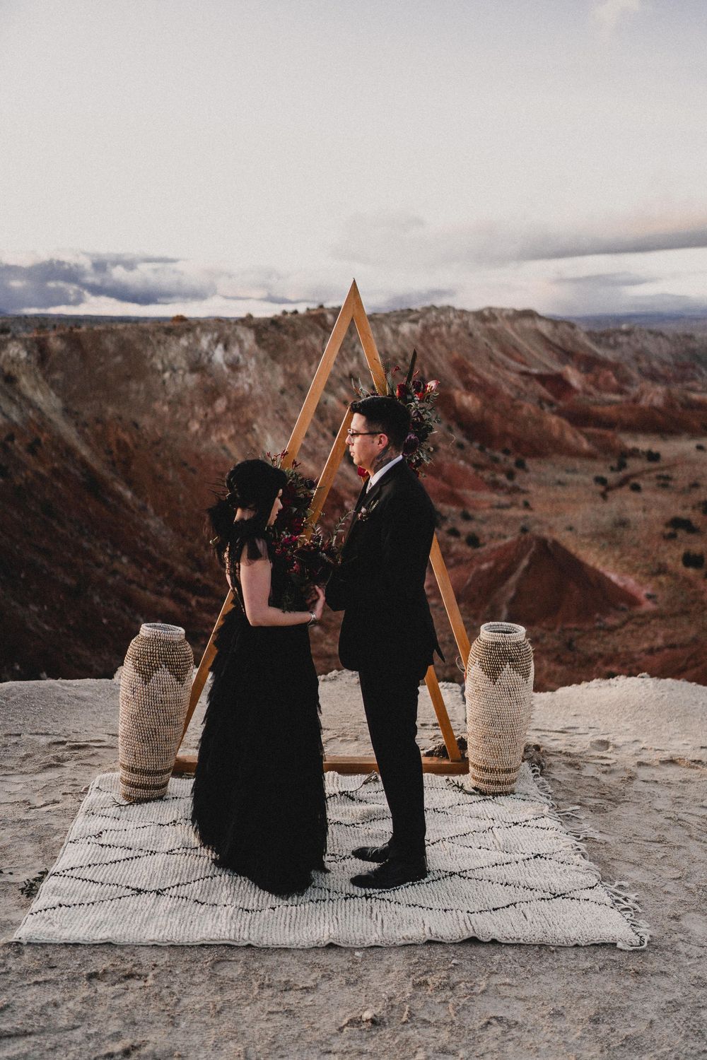 Bride and groom in all-black wedding attire stand at triangular wooden arch over colorful desert canyon in Ojito Wilderness