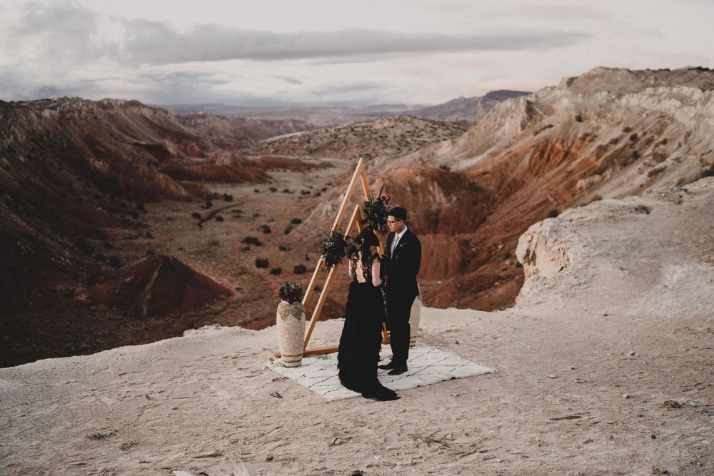 Couple in black wedding attire embrace at triangular arch ceremony setup on dramatic desert canyon rim at Ojito Wilderness
