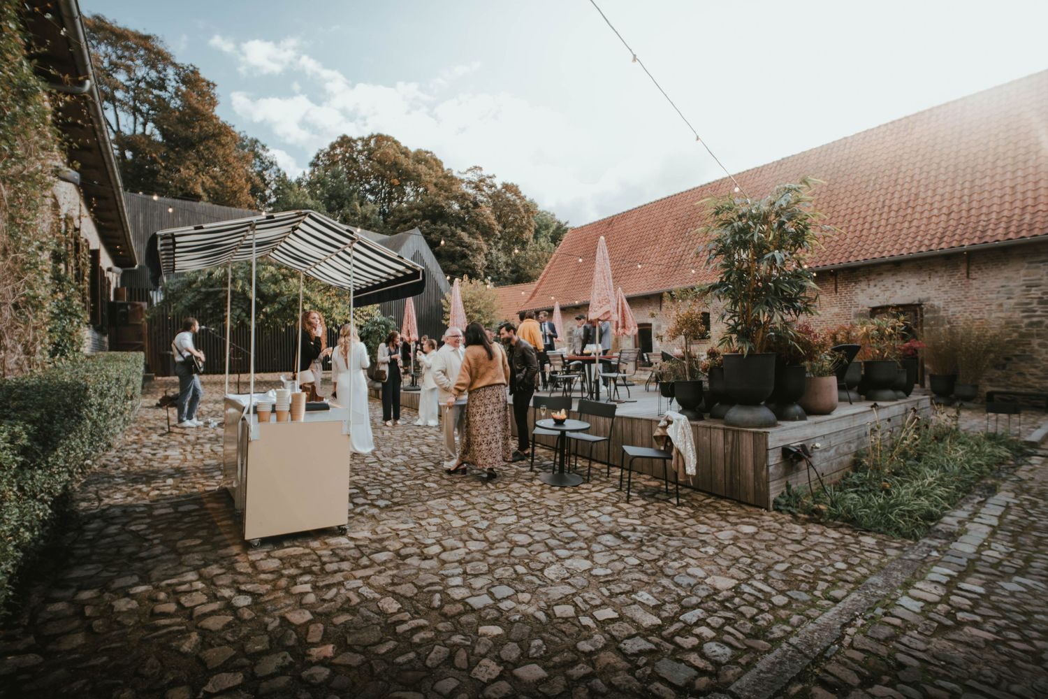 Outdoor wedding reception in a rustic cobblestone courtyard with guests mingling near bar stations and lush greenery.