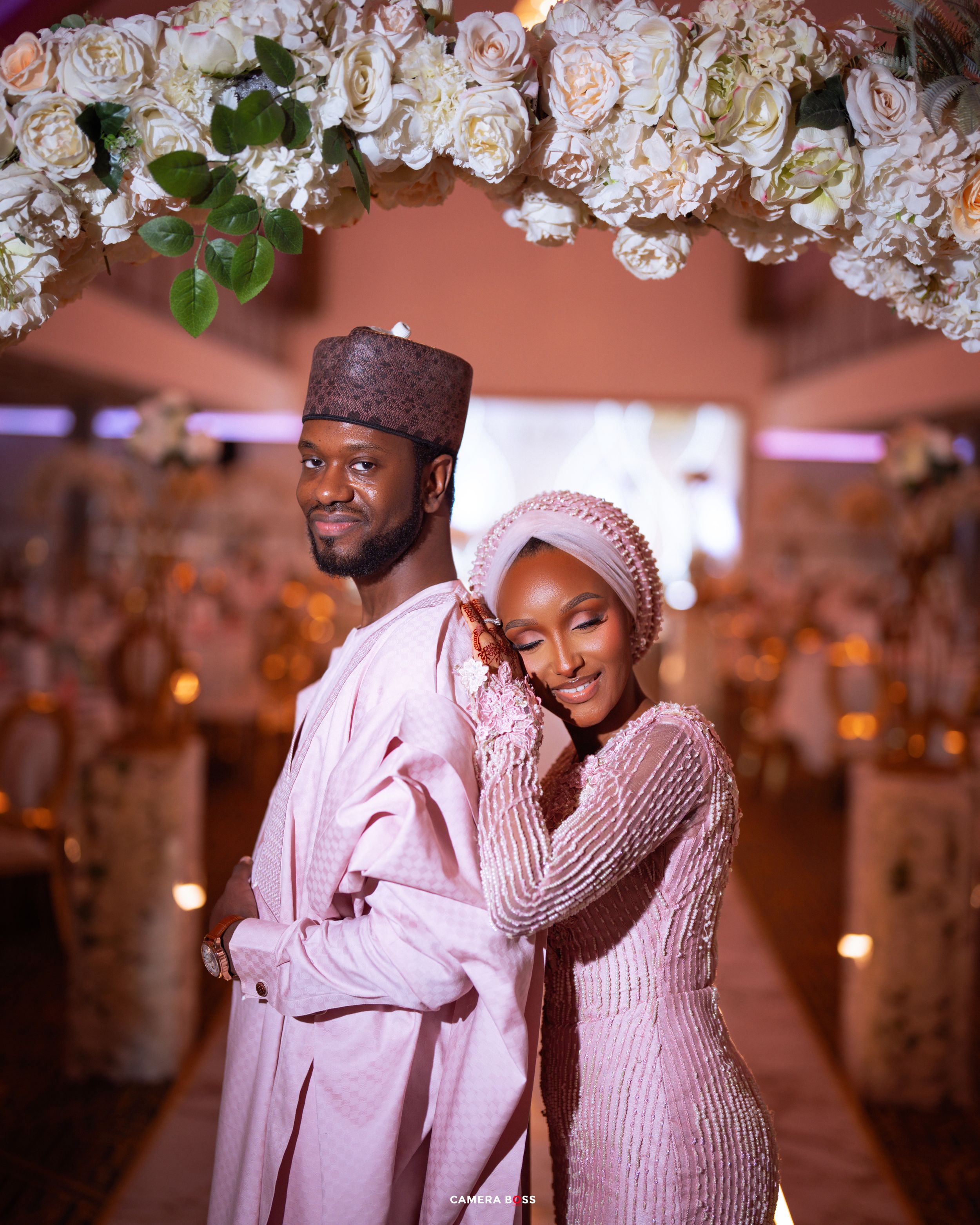 Elegant couple in matching blush pink traditional Nigerian attire posing under floral arch at wedding ceremony.
