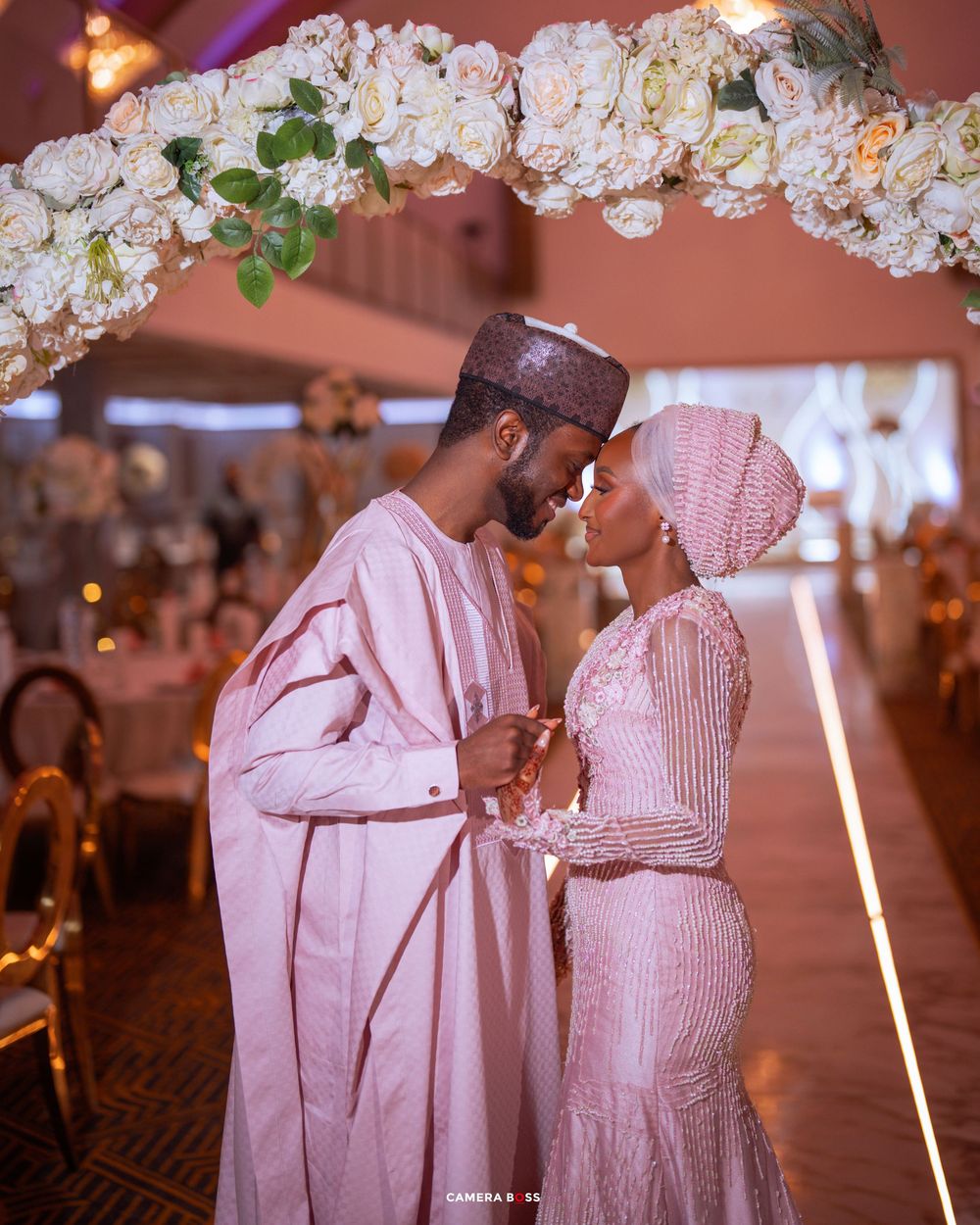 Couple in traditional pink Nigerian wedding attire sharing a romantic moment under a floral arch in an elegant venue.
