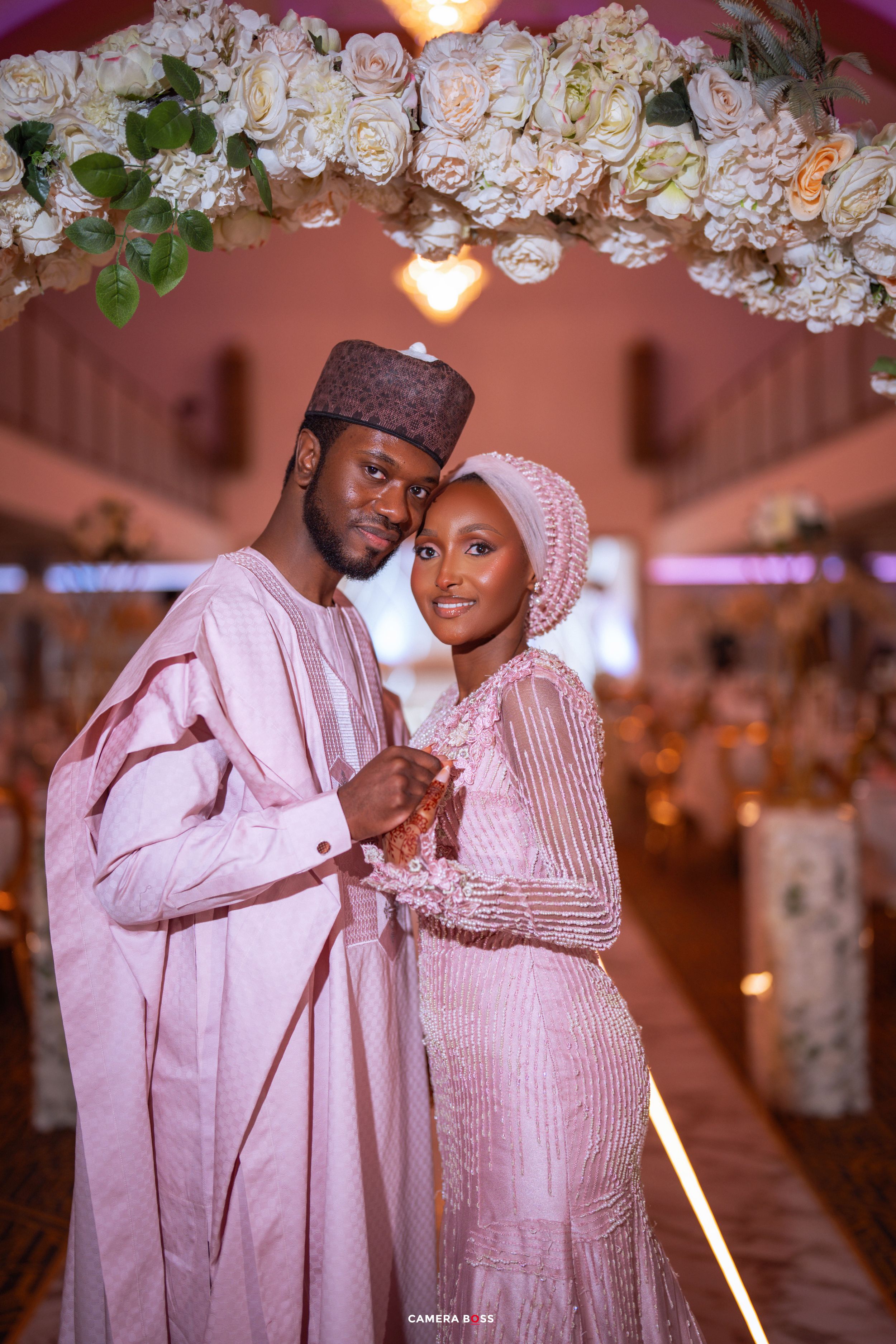 A couple in elegant pink traditional Nigerian attire poses under white floral arch at a wedding reception.