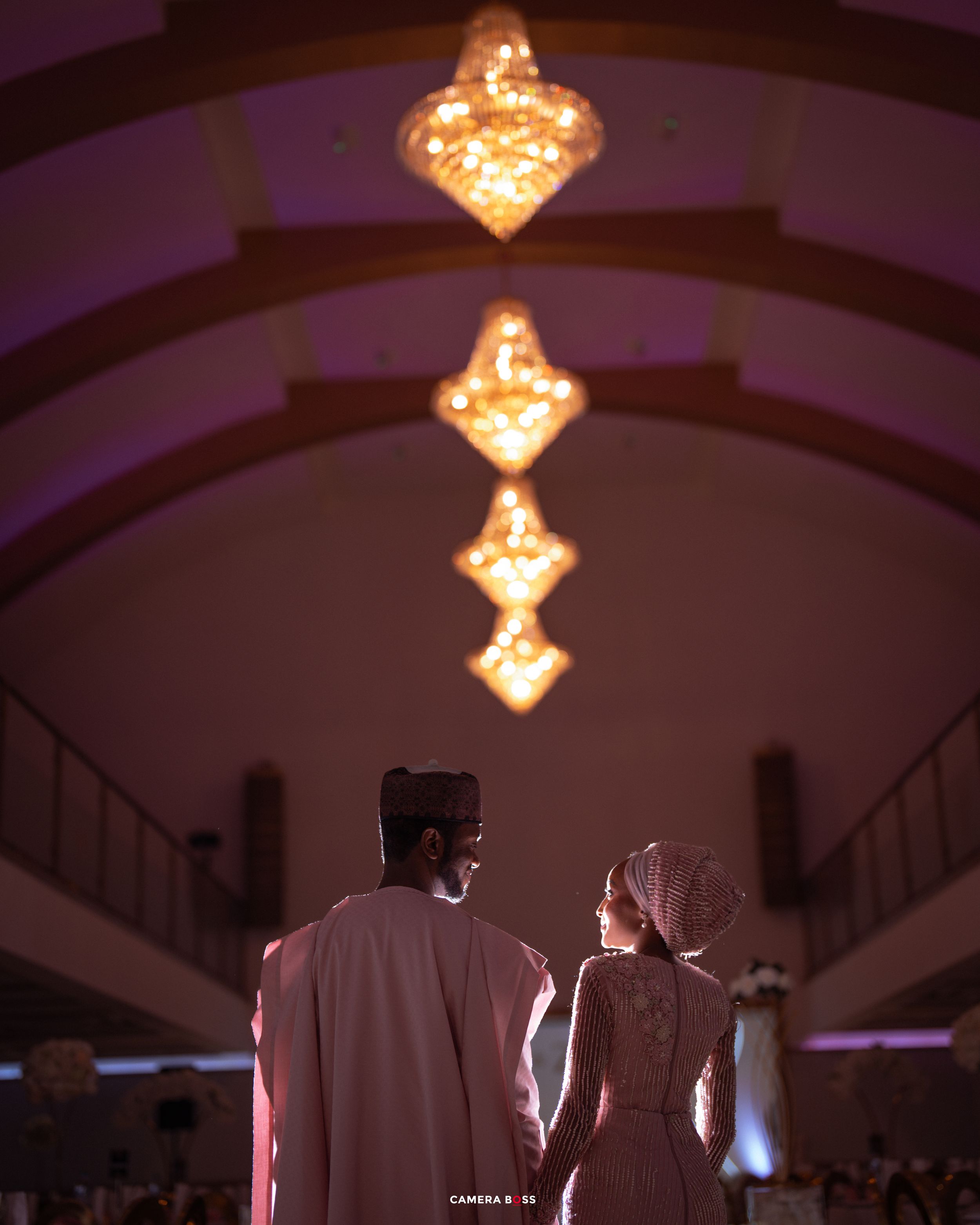 Elegant wedding couple holding hands under chandelier lights in a grand hall with purple ambient lighting.