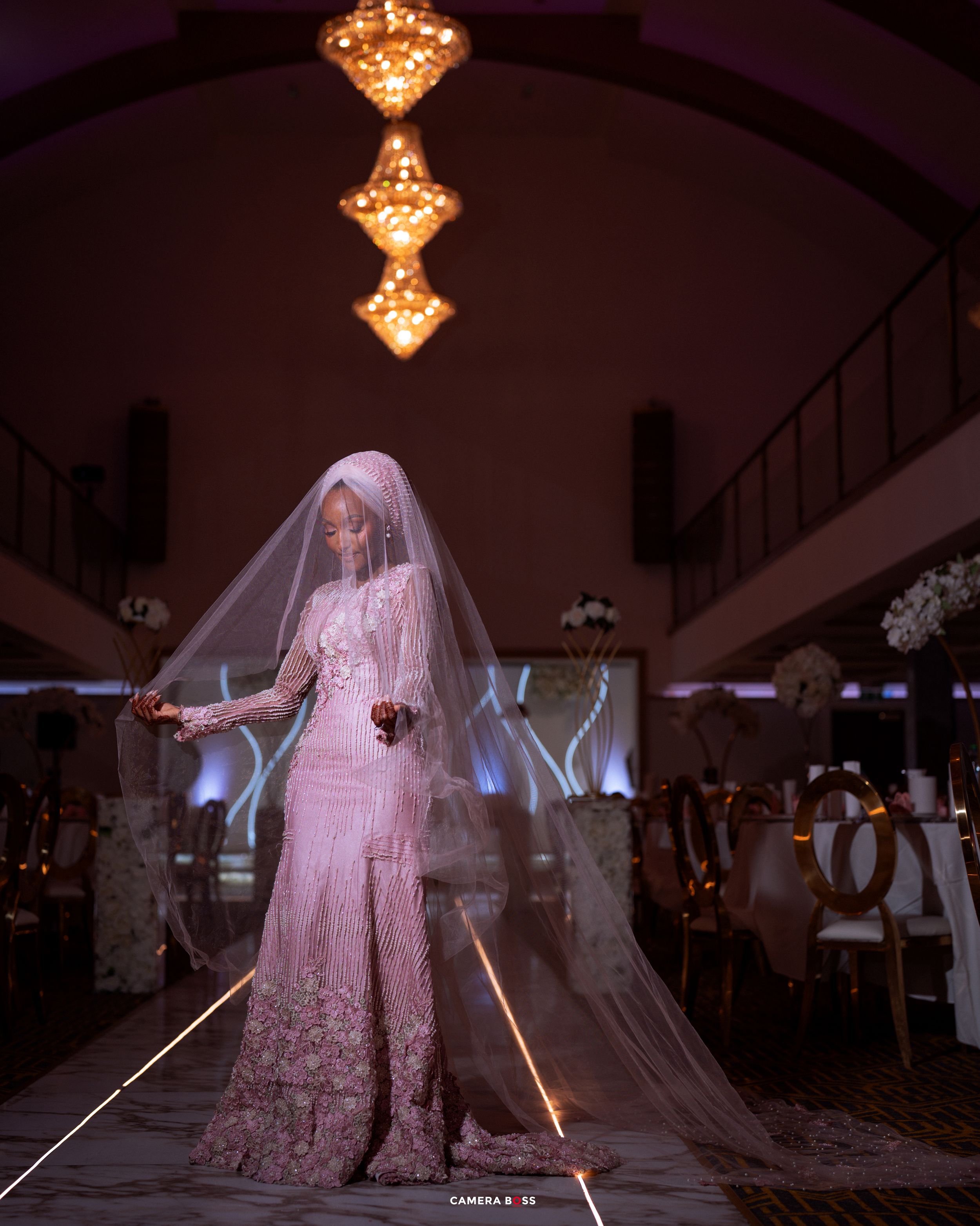 Bride in pink lace gown and long veil stands elegantly in warmly lit wedding venue with chandelier above.